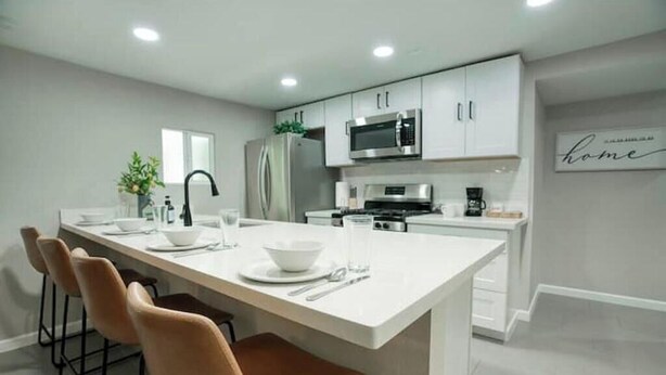 Une cuisine moderne avec des meubles blancs, un réfrigérateur en acier inoxydable, un four à micro-ondes et une machine à café. On y trouve également un coin repas avec une table blanche et des chaises marron.