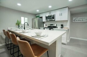 A modern kitchen with white cabinetry, a stainless steel refrigerator, a microwave, and a coffee maker. There is a dining area with a white table and brown chairs.