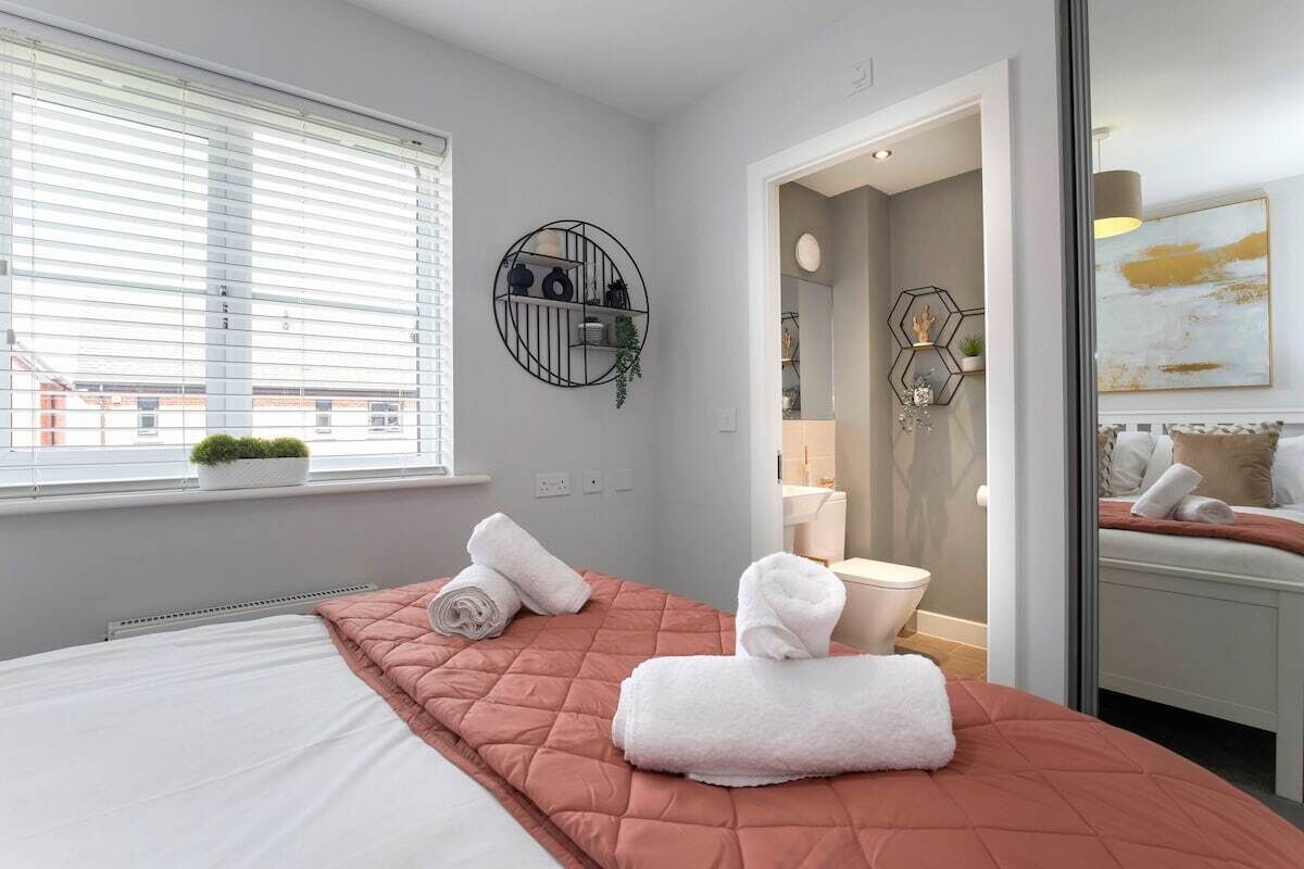Une chambre d’hôtel moderne avec un grand lit, une étagère murale ronde, une fenêtre avec des stores et une salle de bain visible à travers une porte ouverte.