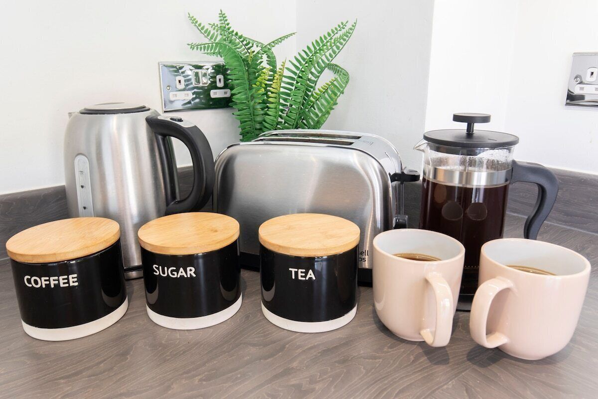 Eine Küchenarbeitsplatte mit Kaffeemaschine, Toaster und einem French Press Kaffeebereiter. Drei beschriftete Behälter für Kaffee, Zucker und Tee sowie zwei Kaffeetassen sind vorhanden.
