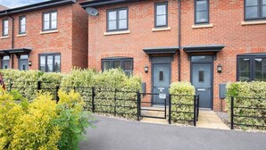 A row of red brick townhouses with black-framed windows and doors, a gated driveway, and a well-maintained hedge. - Modern Two Bedroom Two Bathroom Home (Gloucestershire)