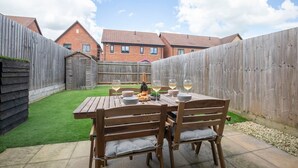 A wooden table with four chairs set outdoors, surrounded by a wooden fence, with a grassy area and a brick building in the background.