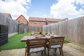 A wooden table with four chairs set outdoors, surrounded by a wooden fence, with a grassy area and a brick building in the background. - Modern Two Bedroom Two Bathroom Home (Gloucestershire)