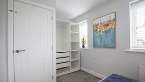 A bedroom with a bed, a white wardrobe, a built-in shelving unit, and a window with blinds. - Modern Two Bedroom Two Bathroom Home (Gloucestershire)