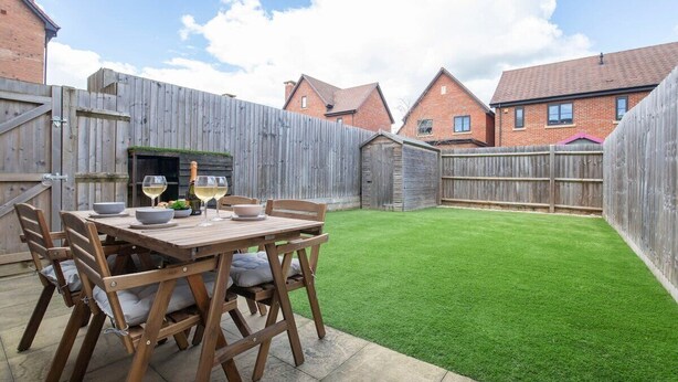 A wooden deck with a table set for two, surrounded by a wooden fence, and a well-maintained lawn. - Modern Two Bedroom Two Bathroom Home (Gloucestershire)