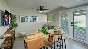 Sala de estar moderna com ventilador de teto, área de jantar com mesa redonda e cadeiras, cozinha visível através de uma porta de vidro e uma janela grande com persianas.