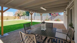 Um pátio externo com mesa e cadeiras, um candeeiro e vista para um quintal com relva e cerca.