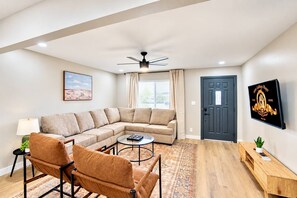 A living room with a sectional sofa, a coffee table, a floor lamp, a television mounted on the wall, and a door.