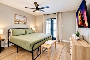 A bedroom with a bed, a bedside table, a nightstand, a ceiling fan, a window with curtains, a wall-mounted TV, and a painting.