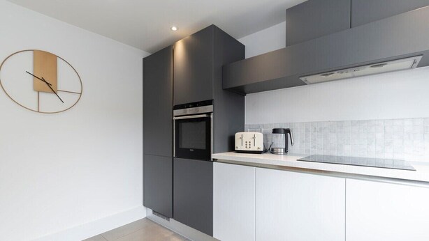A modern kitchen with grey cabinetry, a built-in oven, a coffee maker, and a wall clock. - Peaceful & Spacious 2 Bed Home in Cheltenham (Gloucestershire)