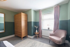 A room with a wooden wardrobe, a pink armchair, and a small table with a vase. - Peaceful & Spacious 2 Bed Home in Cheltenham (Gloucestershire)