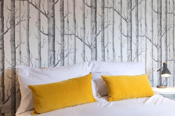 A bed with white bedding and two yellow pillows, set against a wall with a vertical tree pattern wallpaper. - Peaceful & Spacious 2 Bed Home in Cheltenham (Gloucestershire)