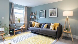 A living room with a grey sofa, yellow cushions, a standing lamp, and a bookshelf. - Peaceful & Spacious 2 Bed Home in Cheltenham (Gloucestershire)