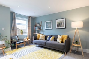 Ein Wohnzimmer mit grauem Sofa, gelben Kissen, einer Stehlampe und einem Bücherregal.