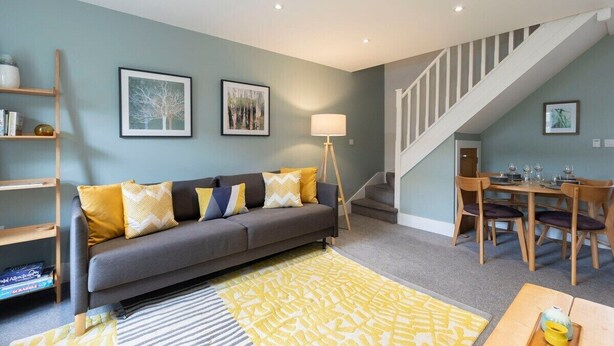 A living room with a grey sofa, yellow cushions, a wooden coffee table, and a staircase leading to a dining area. - Peaceful & Spacious 2 Bed Home in Cheltenham (Gloucestershire)