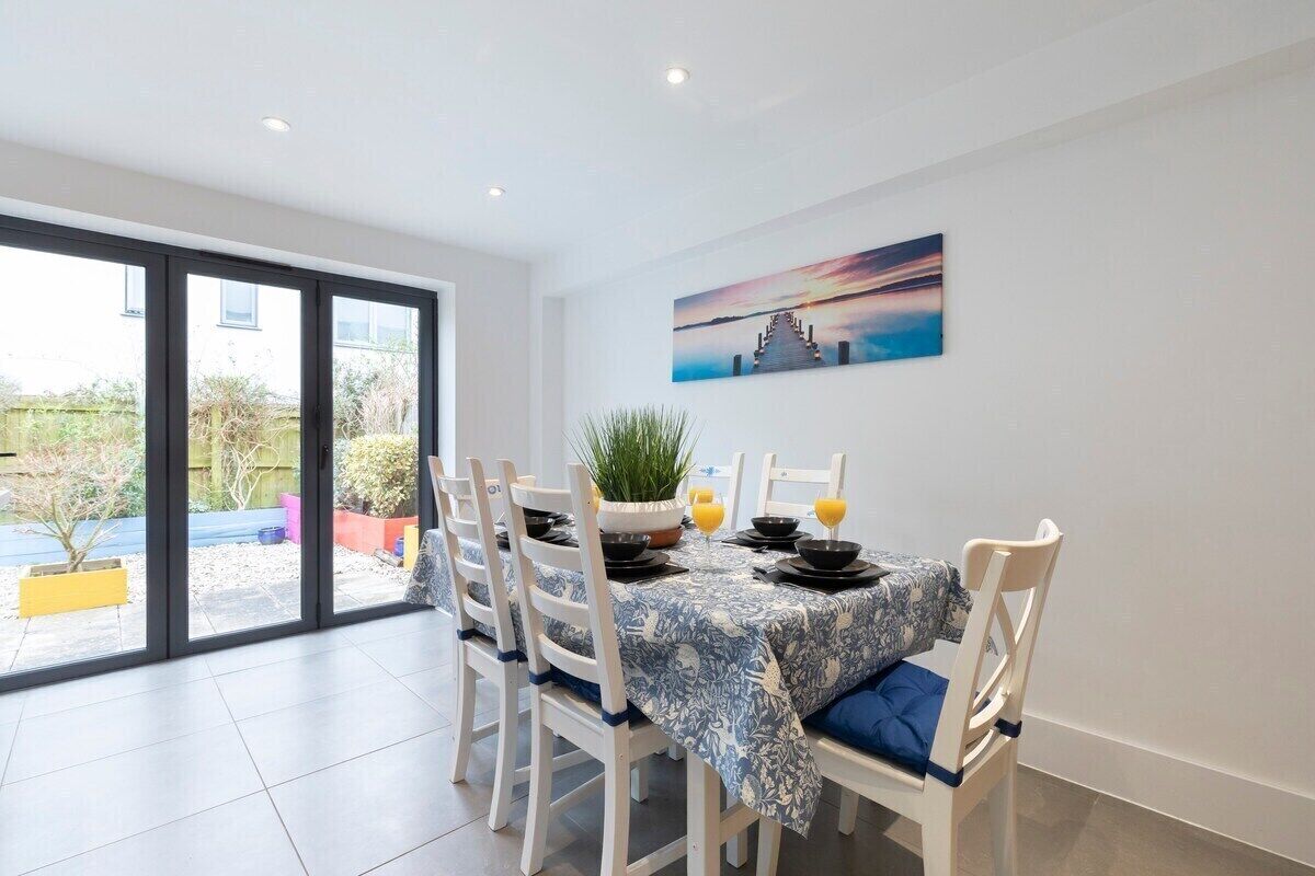 Un comedor con una mesa dispuesta para cuatro personas, sillas blancas, un mantel azul y un cuadro en la pared.