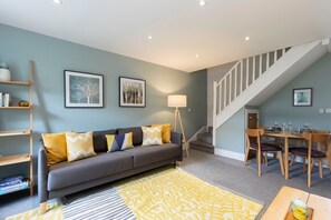 A living room with a grey sofa, yellow cushions, a wooden coffee table, and a staircase leading to a dining area. - Quiet Twin Homes: 5 Beds, 3 Baths & Parking For 3 (Gloucestershire)