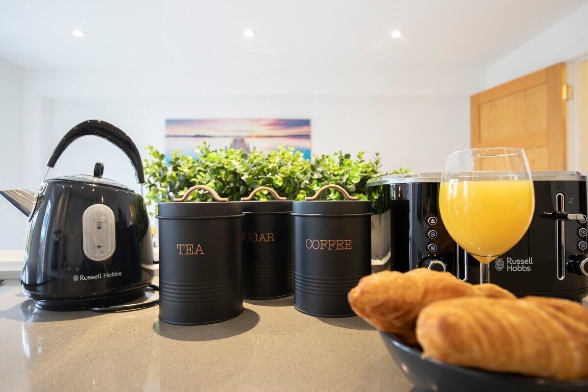 Un plan de travail de cuisine avec une bouilloire, trois récipients étiquetés « thé », « sucre » et « café », un verre de jus d’orange et un bol de croissants.