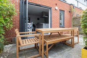 A wooden outdoor table and chairs set against a brick wall, with a garage door open behind. - Quiet Twin Homes: 5 Beds, 3 Baths & Parking For 3 (Gloucestershire)
