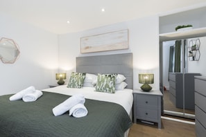 A modern bedroom with a large bed, two bedside tables with lamps, a grey wardrobe, and a mirror.