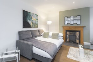 A modern bedroom with a large bed, a grey sofa, a glass coffee table, a painting, a mirror, and a fireplace.