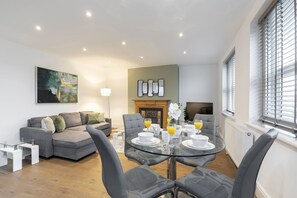 A modern living room with a glass dining table set for four, a grey sofa, and a fireplace.