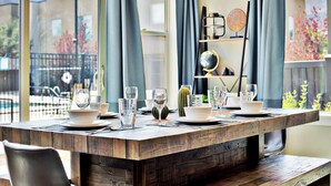A dining table set with plates, glasses, and cutlery, with a view of the outdoors through large windows.