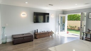 A modern living room with a flat-screen TV, a wooden cabinet, a large vase, and a sliding glass door leading to a garden.