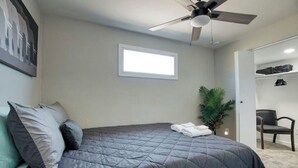 Een slaapkamer met een bed, een plafondventilator, een raam, een plant en een kledingkast met kleding.