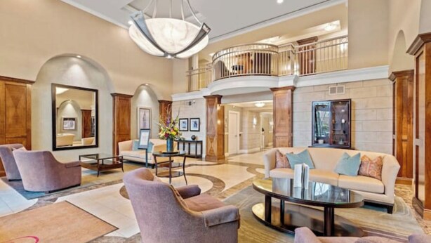 A spacious lobby with a high ceiling, a central seating area with sofas and armchairs, a grand chandelier, and a balcony with a railing.