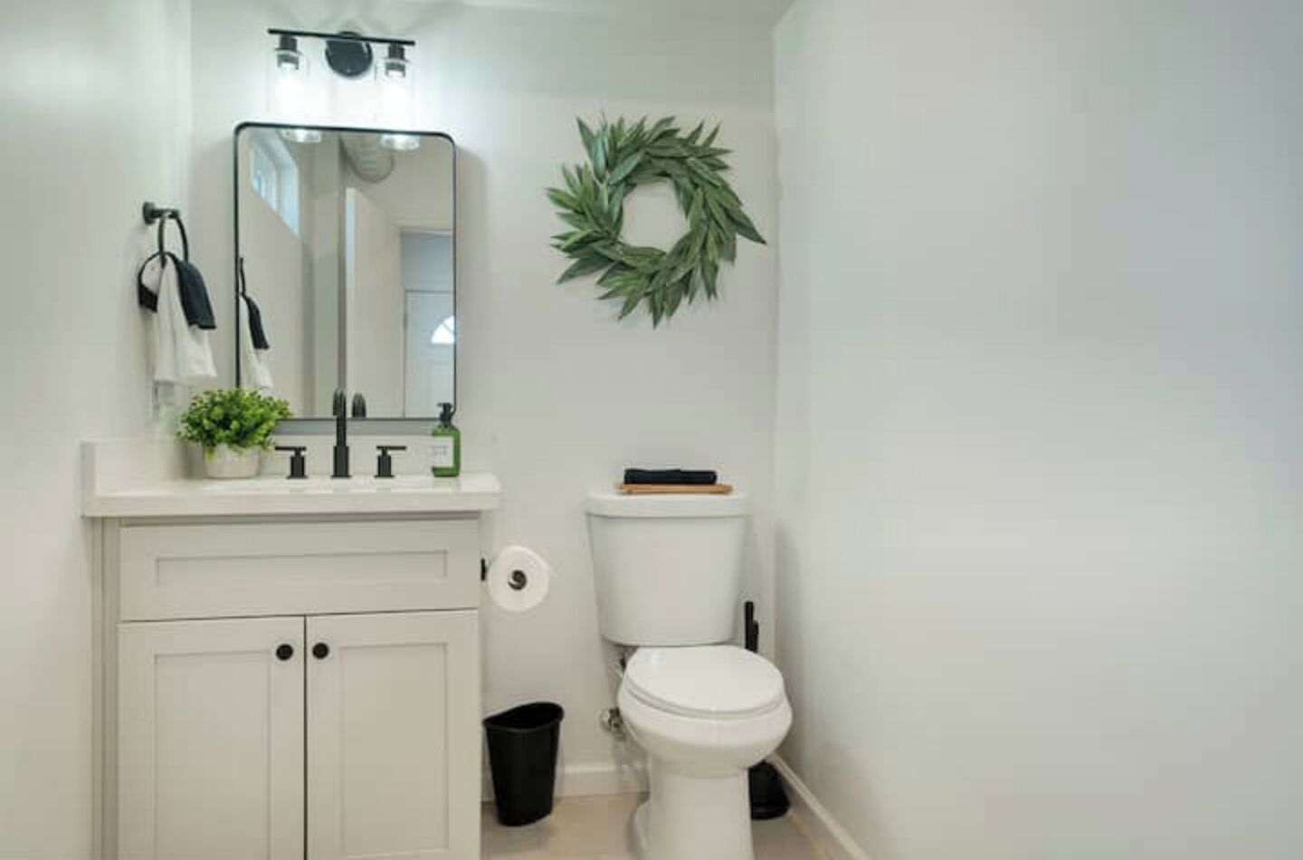Une salle de bain avec un lavabo blanc, un miroir et des toilettes. Il y a une guirlande verte accrochée au mur et une poubelle noire.