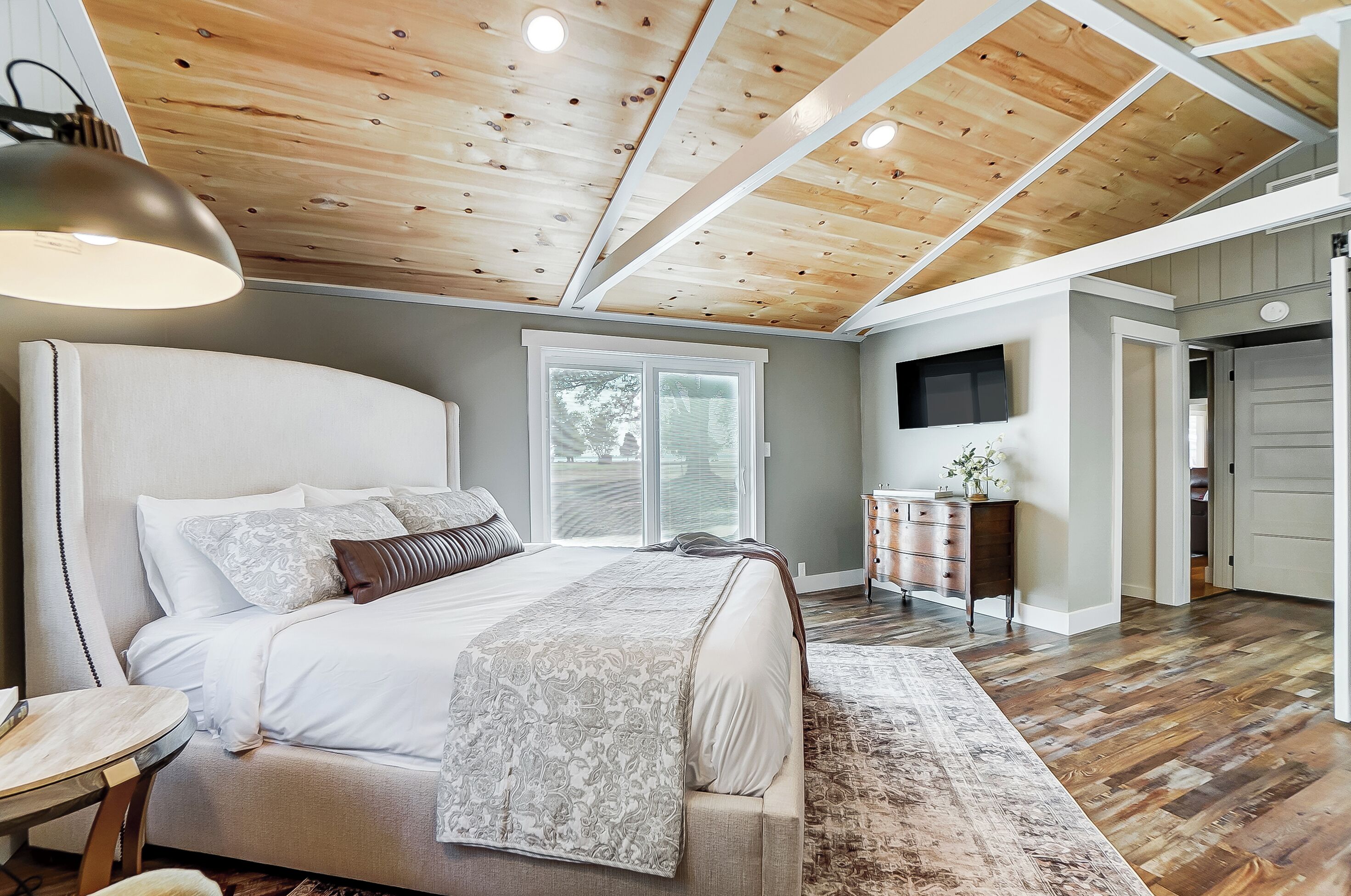 A bedroom with a large bed, a desk, a lamp, a TV, and a wooden ceiling.