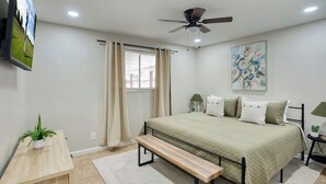 A bedroom with a bed, a bench, a ceiling fan, a window with curtains, a painting on the wall, and a small table with a plant. - Modern Tempe Retreat w/Hot Tub

 (Tempe)
