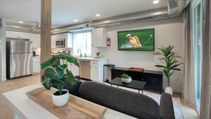 Une cuisine moderne équipée d’appareils électroménagers en acier inoxydable, un coin repas avec une table en bois et un canapé, et un salon doté d’un téléviseur à écran plat diffusant une scène de golf.