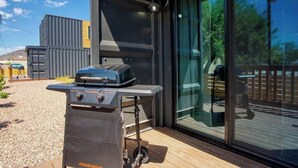 Un espace barbecue extérieur moderne, doté d’un barbecue Megamaster, d’une terrasse en bois et d’une porte vitrée donnant accès à une autre terrasse.