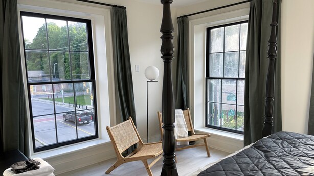 A bedroom with a four-poster bed, two windows with curtains, a chair, and a stack of towels on a nightstand.