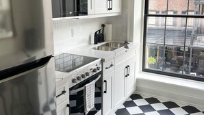A kitchen with white cabinets, a stainless steel refrigerator, a microwave, a stove, and a large window with a view of buildings.