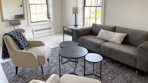 A modern living room with a grey sofa, a round coffee table, and a beige armchair. There are two windows providing natural light, a mirror on the wall, and a small side table with a lamp.