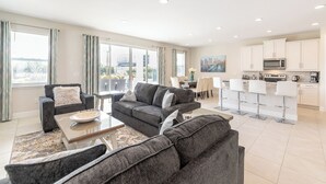 Uma sala de estar moderna com sofás cinza, uma mesa de centro e uma área de jantar com mesa e cadeiras. A cozinha possui armários brancos e eletrodomésticos modernos.