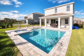 Una piscina con zona de patio, un área de estar cubierta y una casa de dos pisos al fondo.