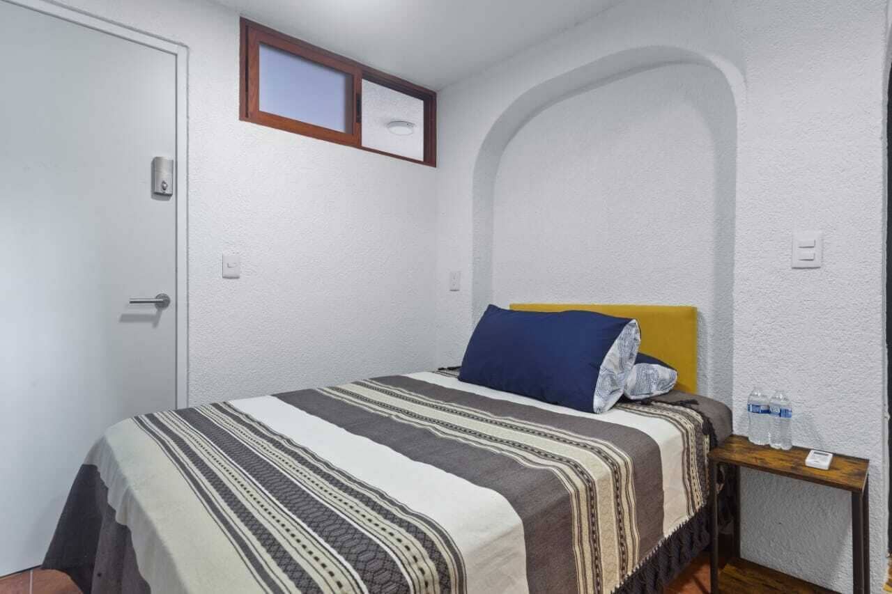 A small, single-bed room with a plain white wall, a wooden-framed picture above the bed, a small table with a water bottle, and a door with a handle.