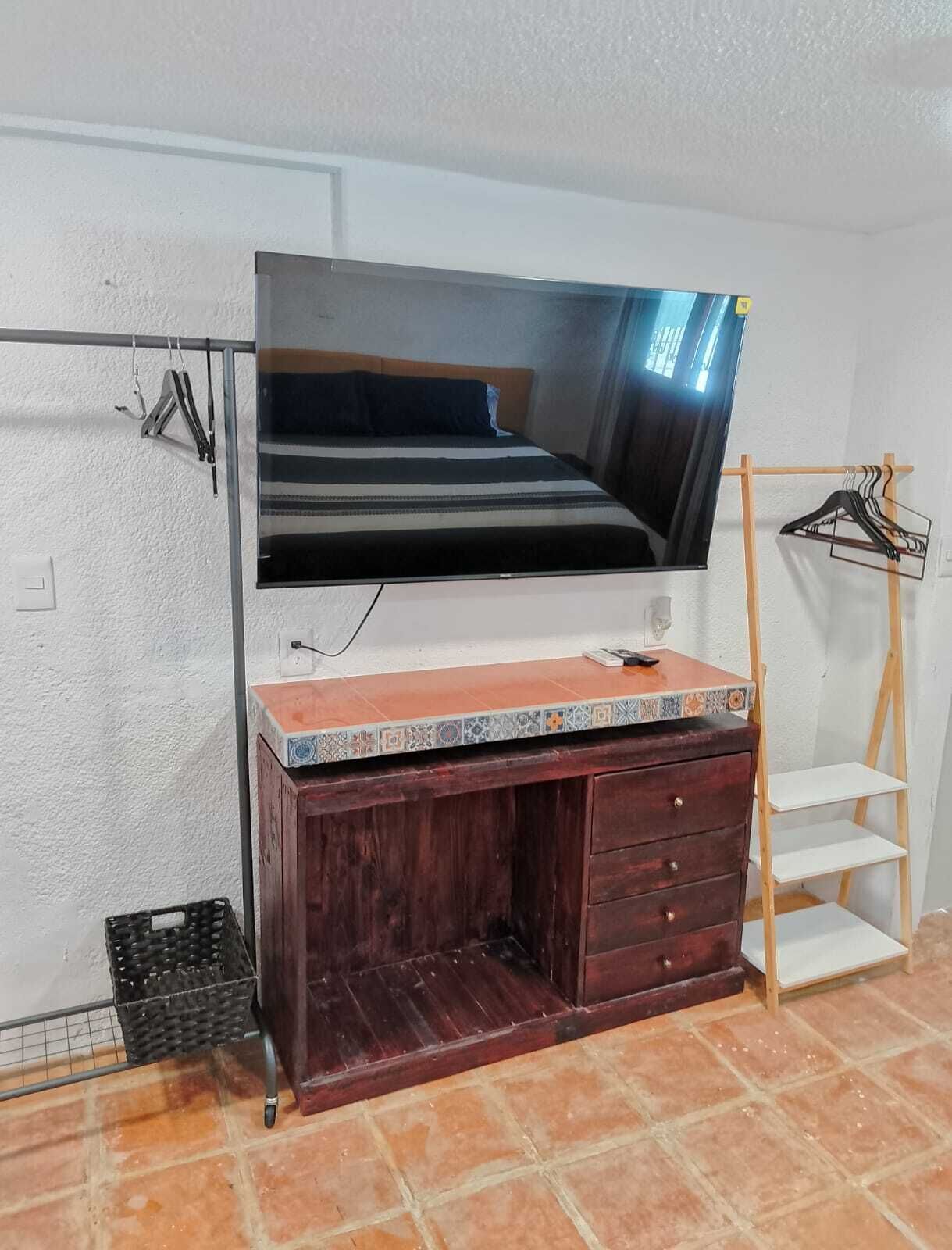 A bedroom with a TV mounted on the wall, a wooden dresser with drawers, a small shelf, and a clothing rack.