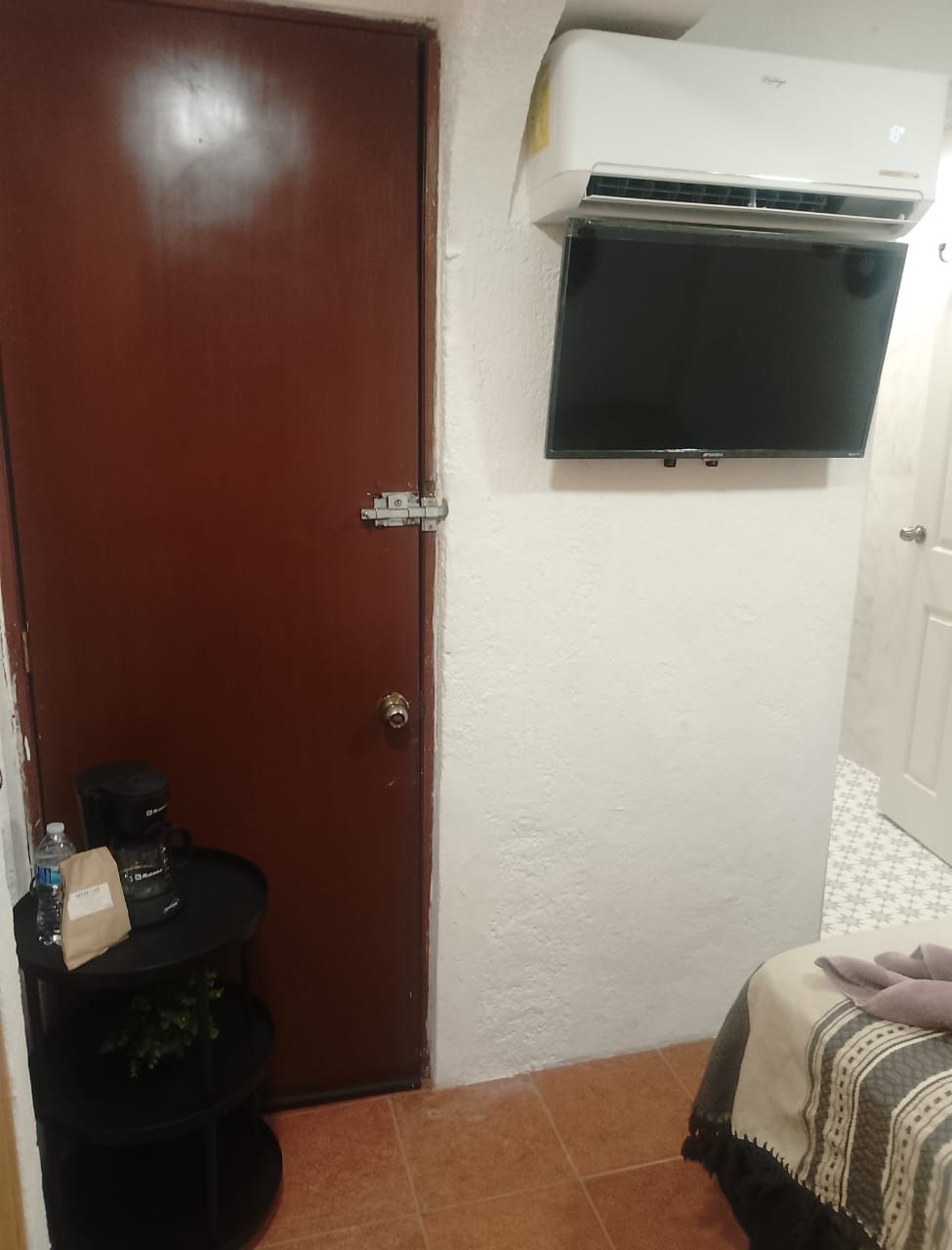 A small room with a wooden door, a black table with a water bottle and a bag, a bed with a blanket, and a mounted air conditioner.