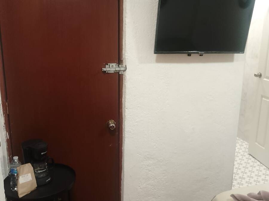 A small room with a wooden door, a black table with a water bottle and a bag, a bed with a blanket, and a mounted air conditioner.
