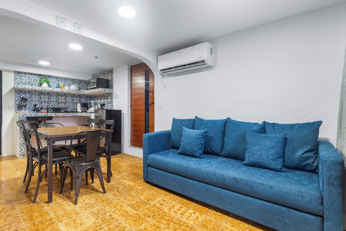 Un espacio habitable compacto con comedor, un sofá azul y una cocina con baldosas estampadas.