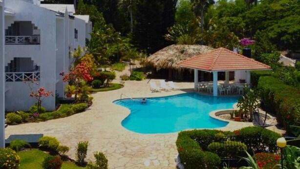 A swimming pool with a thatched-roof pavilion, surrounded by lush greenery and a stone pathway. - Cozy 2-bedroom condo in awesome Sosúa with AC, WiFi, and a gorgeous pool
 (Sosúa)
