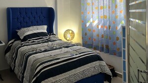 A bedroom with a blue upholstered bed, a bedside table with a lamp, and a window with patterned curtains. - Cozy 2-bedroom condo in awesome Sosúa with AC, WiFi, and a gorgeous pool
 (Sosúa)