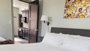 Ein Hotelzimmer mit einem großen Bett, einem Nachttisch, einer Lampe, einem Gemälde an der Wand und Blick auf Küche und Bad durch eine geöffnete Tür.