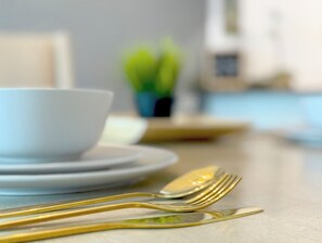 Egy étkezőasztal, rajta egy fehér tál, egy kis virágos növény és arany színű evőeszközök.