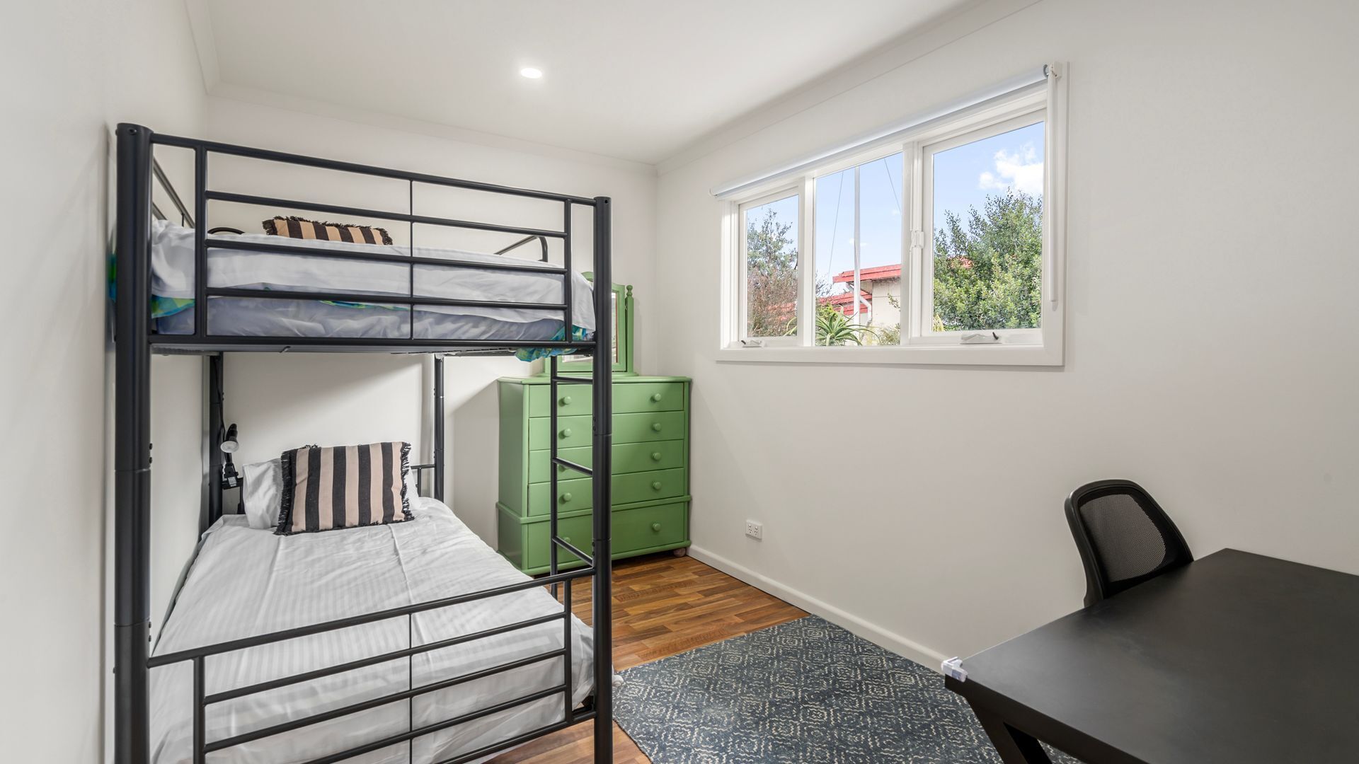 A dormitory room with a bunk bed, a desk, a chair, a dresser, and a window with a view of greenery and a building.